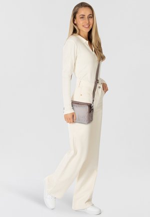 Crossbody bag in textured gray fabric with a zipper closure. Features adjustable strap and embossed logo detail. Model wears cream outfit.