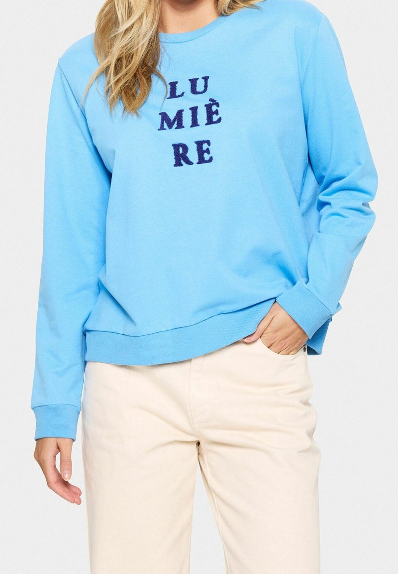 Personne portant une chemise à manches longues bleu clair avec le mot « LUMIÈRE » en lettres bleu marine, associée à un pantalon blanc cassé, main dans la poche.