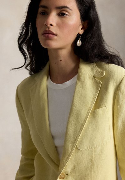 Femme aux longs cheveux foncés portant un blazer jaune clair, un haut blanc et une boucle d'oreille pendante en forme de coquillage à l'oreille gauche.