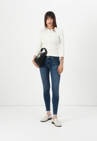 Haut à manches longues blanc avec des boutons, jean skinny bleu foncé, et chaussures slip-on blanches cassées. Sac à main triangulaire noir avec une courte anse.