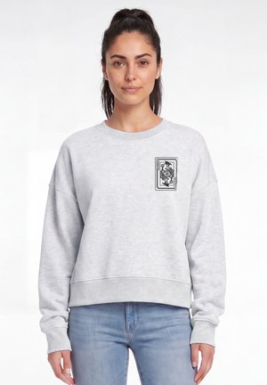 Femme aux cheveux noirs attachés en queue de cheval portant un sweat-shirt gris clair avec un petit motif de carte joker noire sur le haut gauche de la poitrine.