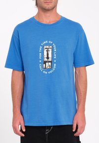 Blå bomulds t-shirt med et centralt grafik af en betalingstelefon og tekst. Korte ærmer, rund hals, afslappet pasform, glat tekstur.