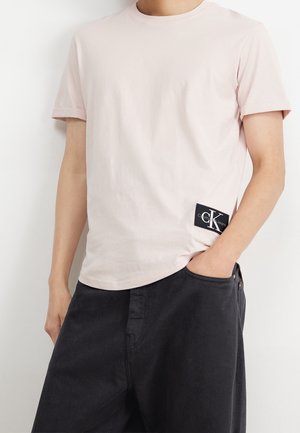Homme portant un t-shirt rose clair à manches courtes avec un patch logo noir Calvin Klein Jeans au-dessus de la poche d’un pantalon noir.