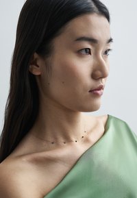 Aranylánc nyaklánc apró fekete drágakövekkel, sima zöld szöveten viselve, kiemelve a minimalista dizájnt.