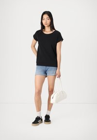 Haut noir à manches courtes, short en denim bleu clair, sac blanc, baskets noires avec accents blancs et chaussettes grises sur un fond minimal.