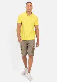 Gelbes Poloshirt mit kurzen Ärmeln, kombiniert mit beigen Cargo-Shorts mit Seitentaschen und kontrastierenden Patches. Weiße Sneakers runden den Look ab.