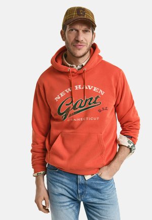 Orange hoodie with a front pocket, featuring white text and a green "Gant" logo. Worn over a patterned shirt, paired with blue denim jeans.