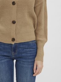 Bežový pletený kardigán s veľkými gombíkmi, nosený cez modré džíny, ukazujúci uvoľnený strih a detail rúk odpočívajúcich po boku.