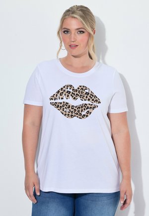 Frau mit blonden Haaren, die Creolen trägt und ein weißes T-Shirt mit großem Leoparden-Lippen-Design, stehend vor hellem Hintergrund.