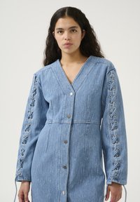 Denim haljina svijetloplave boje s V-izrezom, gumba sprijeda i vezanjem na rukavima, s teksturiranim završetkom i tijesnim strukom.