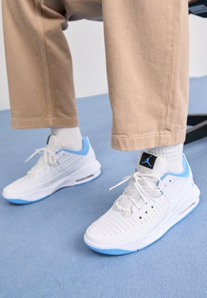 Person iført hvide og lyseblå sneakers med hvide sokker og beige bukser, stående på en blå tæppebelægning.