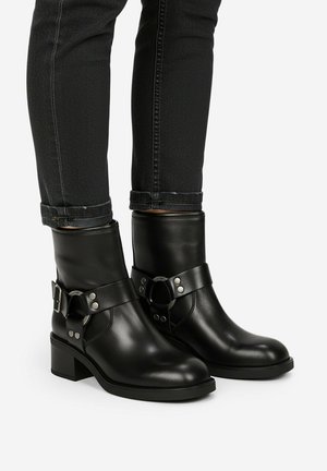 Czarne skórzane botki za kostkę z paskami z srebrnymi klamrami, noszone z czarnymi podwiniętymi obcisłymi dżinsami na jasnym tle.