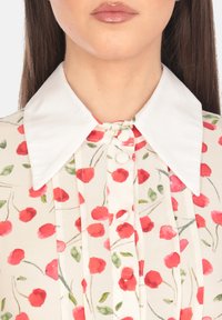 Blusa floreale con colletto bianco, caratterizzata da motivi di rose rosse su uno sfondo crema, tasca con bottoni e pieghe verticali.