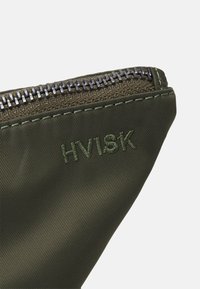 Gros plan sur le coin d'une pochette en tissu vert olive avec une fermeture éclair en métal et le mot « HVISK » brodé en fil vert assorti.