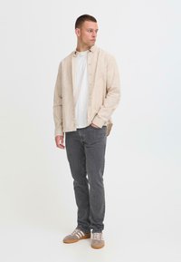 Beige gestreept overhemd met knopen over een wit T-shirt, gecombineerd met grijze spijkerbroek en grijze sneakers. Eenvoudig ontwerp, casual pasvorm, geen zichtbare logo's.