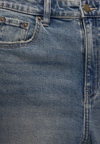 Gros plan sur un jean en denim bleu montrant la poche avant, la petite poche à monnaie avec rivets métalliques, la passants de ceinture et le bouton supérieur avec des coutures visibles.