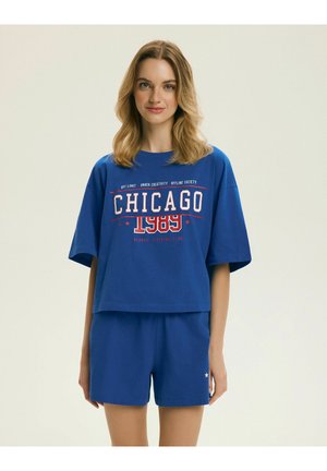 Mladá žena so svetlými vlasmi, ktorá má na sebe modré oversized tričko Chicago 1989 a zodpovedajúce šortky na jednoduchom pozadí.
