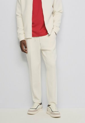 Persona con pantalones deportivos blancos, chaqueta blanca con cremallera a juego, camiseta roja y zapatillas blancas con detalles en beige y negro.