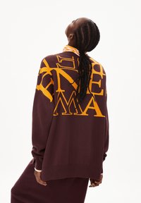 Burgunderfärgad oversized tröja med stor gul grafisk text. Mjuk stickad kvalitet, ribbade ärmslut och nederkant, samt avslappnad passform.