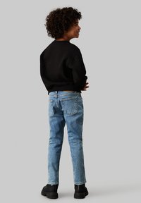 Bambino che indossa un felpa nera a coste e jeans in denim blu chiaro. Stivali neri visibili. La vista da dietro mette in evidenza il taglio casual e il design.