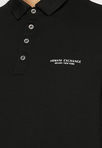 Svart polotröja i bomull, med klassisk krage, fem knappar och en vit broderad "Armani Exchange"-logotyp på bröstet.