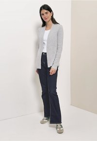 Harmaa resorineuletakki, jossa avoin etuosa, päällä valkoinen toppi, yhdistetty tummansinisiin farkkuihin ja vihrein yksityiskohdin varustettuihin lenkkareihin, seisomassa neutraalia taustaa vasten.