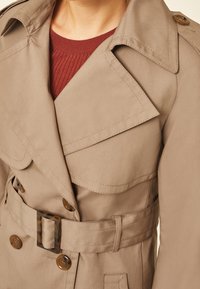 Gros plan d'un trench beige avec larges revers, détails de boutons et ceinture, porté sur un pull rouge côtelé.