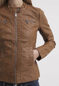 Brun läderjacka med ståkrage, framdragkedja och två fickor med dragkedja på bröstet. Har en strukturerad yta och figurnära passform.