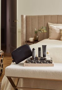 Svart toalettmappe med glidelås og "Rituals"-logo, omringet av forskjellige hudpleieprodukter som er vist på et mønstret brett oppå en teksturert overflate.