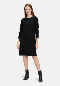 Robe noire à manches longues avec un col rond, texture unie, coupe mi-longue et poignets côtelés, associée à des bottes noires hauteur cheville.