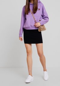 Fialová mikina s kapsou na přední straně, zkombinovaná s černou mini sukní. Bílé tenisky a malá texturovaná kabelka dotvářejí outfit.