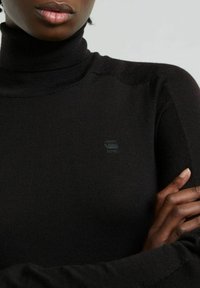 Maglione a collo alto nero realizzato in tessuto a costine, con un logo discreto sul petto e un design aderente con maniche lunghe.