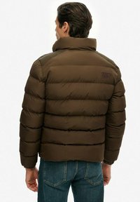 Mand iført en brun polstret jakke med høj krave og blå jeans, vist fra ryggen mod en ensfarvet baggrund.