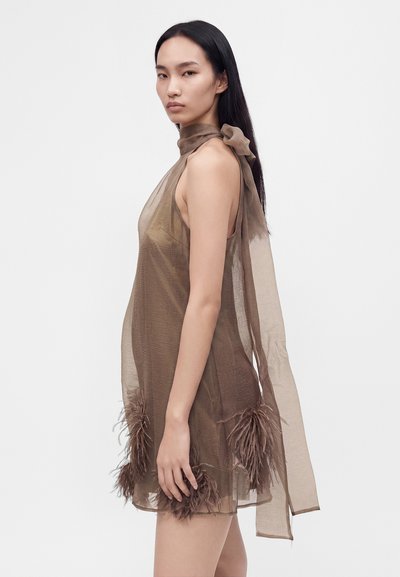 Femme portant une robe marron transparente sans manches avec des détails en plumes à l'ourlet et une longue écharpe nouée autour du cou sur un fond blanc uni.