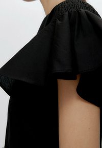 Haut noir avec un décolleté smocké et de grandes manches froncées. Le tissu a une texture lisse, mettant en valeur le design volumineux des manches.