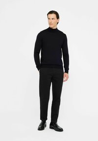Maglione nero a collo alto realizzato in un tessuto jersey liscio ed elasticizzato. Accompagnato da pantaloni neri e scarpe eleganti nere lucide. Design semplice e dalle linee aderenti.