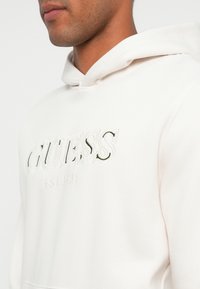 Sudadera blanca con el logo en relieve "GUESS" y acentos verdes, además del texto "EST 1981". Capucha ajustable con cordones y tela suave y lisa.