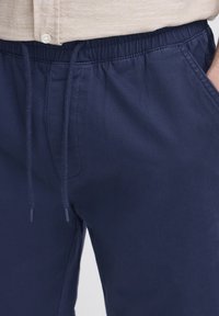 Tamsiai mėlyni šortai iš minkštos medžiagos su elastingu liemeniu ir raišteliu. Matomos šoninės kišenės, su lygiu paviršiumi.