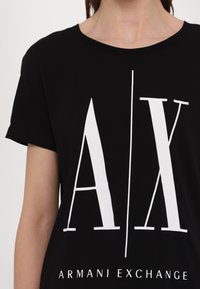 Čierne tričko s krátkym rukávom vyrobené z bavlny, na prednej strane s veľkým bielym logom "A/X" a textom "ARMANI EXCHANGE" na spodnej časti. Hladká textura.