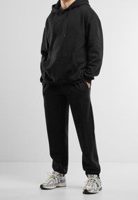 Person trägt einen schwarzen Kapuzenpullover und schwarze Jogginghosen mit silbernen und weißen Laufschuhen, steht vor einem einfarbigen hellen Hintergrund.