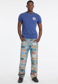 Młody mężczyzna w niebieskiej koszulce i jasnych dżinsach z kolorowymi, markowymi naszywkami, stojący z rękami w kieszeniach.