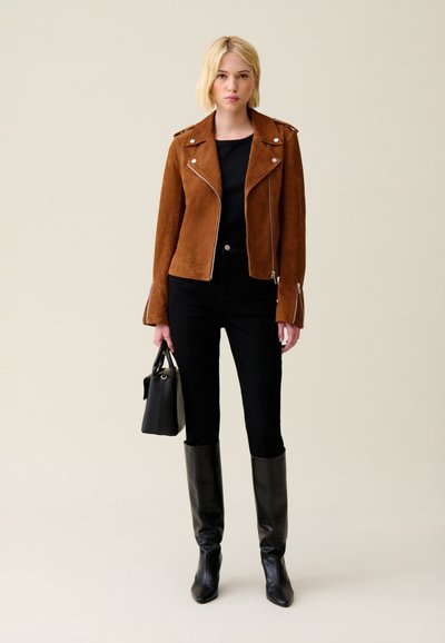 Giacca da moto in suede marrone con cerniere argentate, top nero, jeans neri e stivali neri al ginocchio. Tiene in mano una piccola borsa nera.