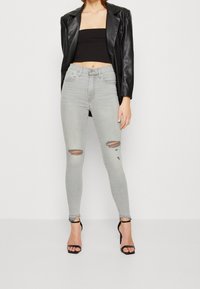 Mujer vistiendo pantalones pitillo rasgados de color gris claro, top corto negro, blazer de cuero negro y sandalias de tacón alto negras, de pie contra un fondo liso.