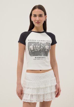 Junge Frau trägt ein schwarz-weißes Ramones-Grafik-T-Shirt und einen weißen mehrlagigen Spitzen-Minirock, steht vor einem schlichten Hintergrund.