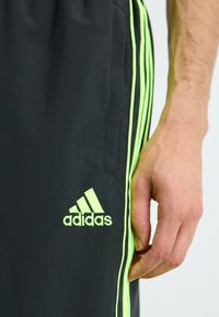 Kadar izbliza ruke pored crnih Adidas hlača s neonsko zelenim prugama i logotipom na bedru, protiv jednostavne pozadine.