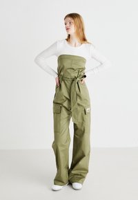 Oliven grønn jumpsuit med cargo-lommer, justerbart knytebelte i midjen og brede ben, lagd over en tettsittende hvit langermet topp.