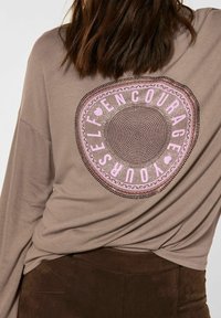 Langärmliges taupefarbenes Oberteil mit einem zirkulären Design auf dem Rücken, das den Text "ERWIESE DICH SELBST" in Pink zeigt, verziert mit Funkeln.
