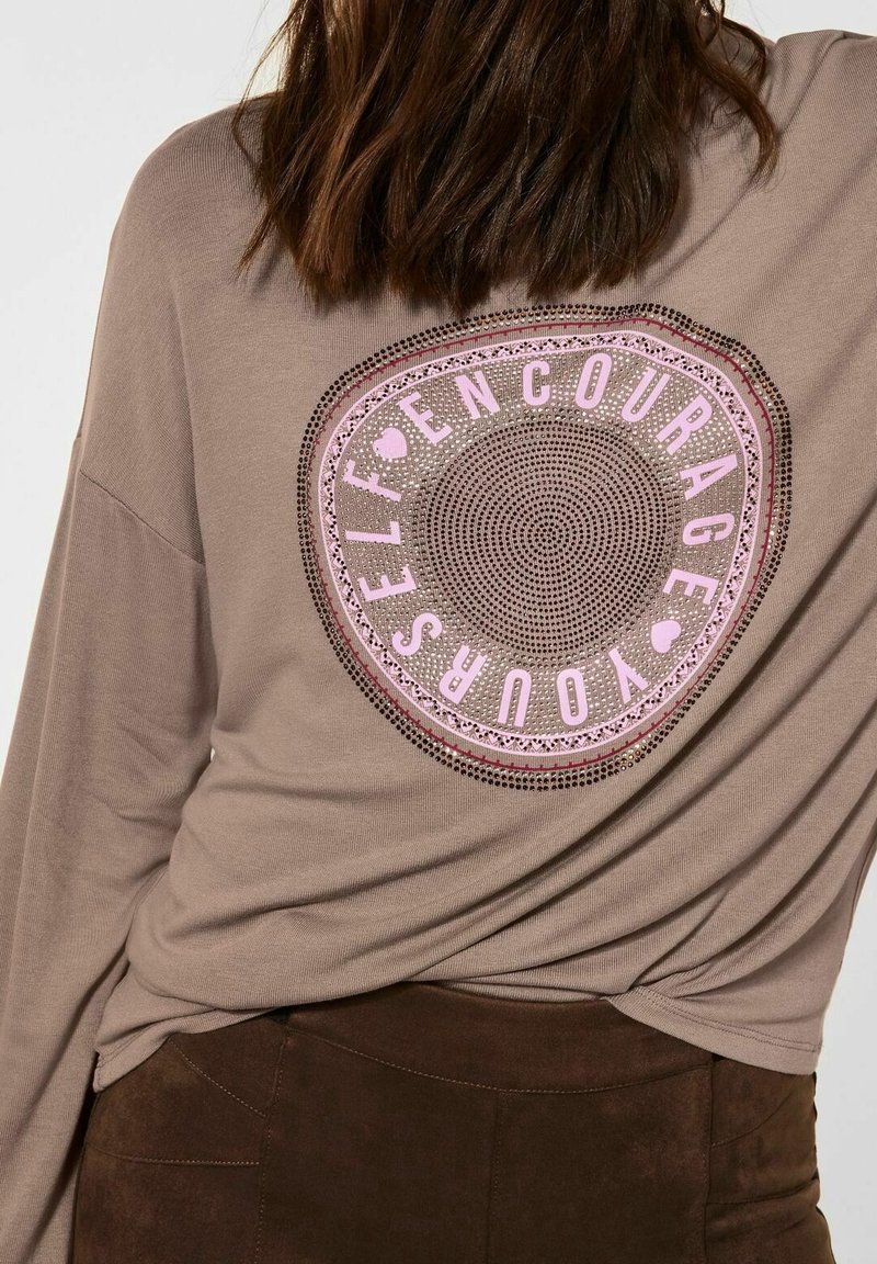 Langärmliges taupefarbenes Oberteil mit einem zirkulären Design auf dem Rücken, das den Text "ERWIESE DICH SELBST" in Pink zeigt, verziert mit Funkeln.