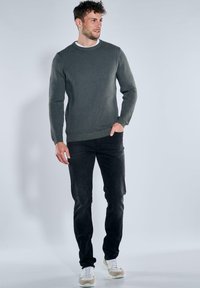 Dunkelgrüner, gerippter Pullover mit Rundhalsausschnitt, kombiniert mit schwarzen, schmal geschnittenen Jeans und hellen Sneakers. Glatte Textur und figurschmeichelndes Design.