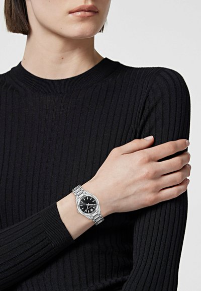 Personne portant un haut noir à manches longues côtelé avec une montre-bracelet en métal argenté avec un cadran noir.